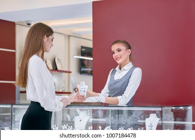 Beautiful Jewelry Store Worker Wearing White Blouse And Grey Uniform Is Showing A Necklace Designed With Blue Stones To A Smartly Dressed Female Client Of The Modern Jewelry Shop.