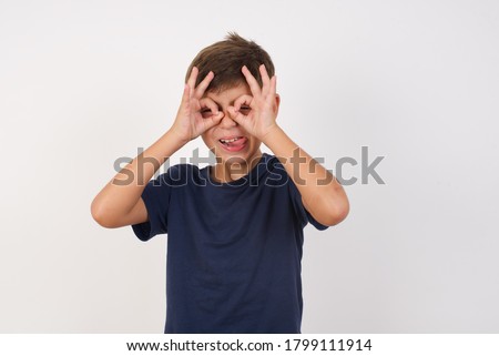 Beautiful kid boy wearing casual t-shirt standing over isolated white background doing ok gesture like binoculars sticking tongue out, eyes looking through fingers. Crazy expression.