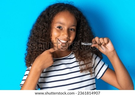 beautiful kid girl with afro curly hairstyle wearing striped T-shirt holding an invisible aligner and pointing to her perfect straight teeth. Dental healthcare and confidence concept.