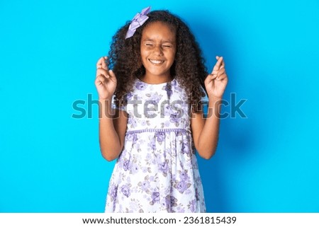 beautiful kid girl with afro curly hairstyle wearing flowered dress has big hope, crosses fingers, believes in good fortune, smiles broadly. People and wish concept