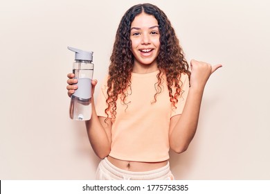 Beautiful Kid Girl With Curly Hair Wearing Sportswear Holding Water Bottle Pointing Thumb Up To The Side Smiling Happy With Open Mouth 