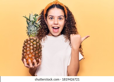 Beautiful Kid Girl With Curly Hair Holding Pineapple Pointing Thumb Up To The Side Smiling Happy With Open Mouth 