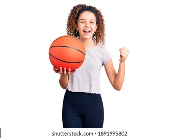 Beautiful Kid Girl With Curly Hair Holding Basketball Ball Screaming Proud, Celebrating Victory And Success Very Excited With Raised Arms 