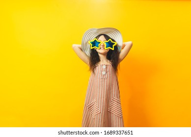 Beautiful Kid Girl In Stripped Dress, Summer Big Hat And Yellow Star Shaped Big Sunglasses Posing With Arms Raised. Three Quarter Length Studio Shot On Yellow Background. Summer Celebration Concept.