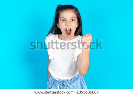 Beautiful kid girl wearing white T-shirt over blue background angry and mad raising fist frustrated and furious while shouting with anger. Rage and aggressive concept.