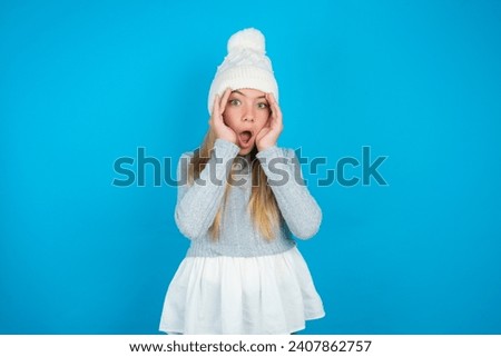 Beautiful kid girl wearing white knitted hat and blue sweater with scared expression, keeps hands on head, jaw dropped, has terrific expression. Omg concept
