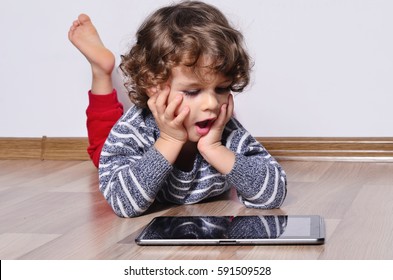 Beautiful Kid Playing Games On A Tablet. Amazed Boy Sitting On The Floor And Looking On The Display Of A Tablet Watching Cartoons.