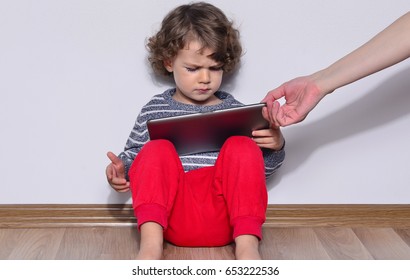 Beautiful kid playing games on a tablet. Boy sitting on the floor and looking on the display, screen of a tablet watching cartoons. Very angry kid because his mother took the tablet from him. 