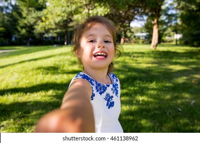 Beautiful Kid Taking Selfie In Green Summer Park, Laughing, Having Fun. Warm Sunny Weather, Healthy Child, Happiness, Freedom, Emotions. Cute Girl Reaching Hand To Camera. 