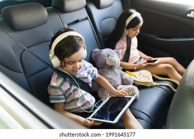 Beautiful Kids Watching A Movie On A Tablet With Headphones While Sitting In The Car. Little Siblings Enjoying Going With Their Parents On A Trip  