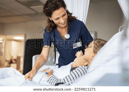 Beautiful kind female nurse taking care of little boy hospitalized in bed. Happy woman nurse tuck the covers back to the young child patient lying on hospital bed. Medical worker with kid patient.