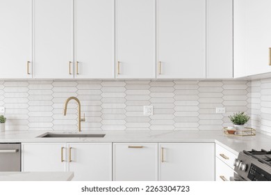 A beautiful kitchen faucet detail with white cabinets, a gold faucet, white marble countertops, and a brown picket ceramic tile backsplash. - Shutterstock ID 2263304723