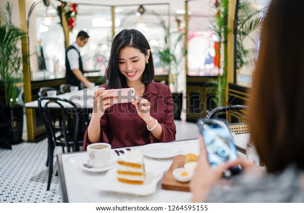A beautiful Korean Asian woman is taking a photograph of\
hear food before having tea and cake with a friend in a trendy\
cafe. 
