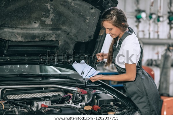 A\
beautiful lady in a black jumpsuit and a white t-shirt is smiling,\
checking the oil level in a black car in the\
garage.
