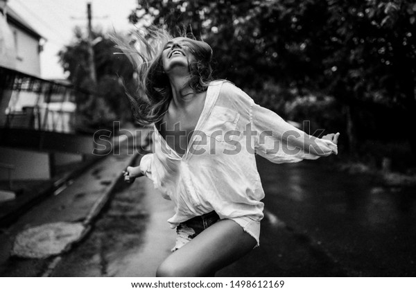 Beautiful lady enjoys fresh rai, she walking\
outdoor in wet clothes and laughing. White big shirt on her sexy\
body, short jeans. Tanned soft skin, long hair, white smile. Trees\
and road behind