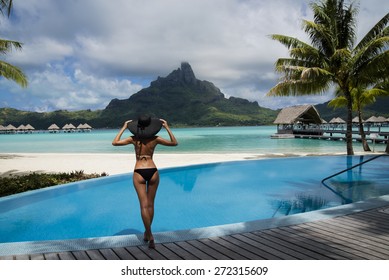 Beautiful Lady In Hat Standing On The Edge Of In The Infinity Pool Looking Through The Lagoon On The Otemanu Mountain On The Tropical Island Of Bora Bora, Near Tahiti, French Polynesia, Pacific Ocean