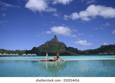 Beautiful Lady Laying On The Edge Of Infinity Pool Of Luxury Resort On Beach With View On  Otemanu Mountain Through Turquoise Lagoon On The Tropical Bora Bora Island, French Polynesia, Pacific Ocean