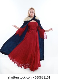 Beautiful Lady With Long Blonde Hair Wearing A Red Medieval Fantasy Gown And A Blue Fur Cloak. Standing, Isolated On White Background.