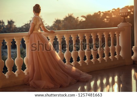 Beautiful lady in luxurious ballroom dress with tulle skirt and lacy top standing on the large balcony looking away at sunset. Back view. Text space