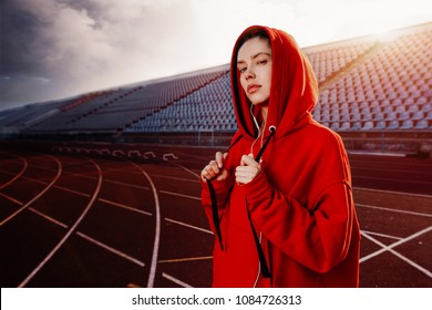 Beautiful lady runner in warm clothes on the stadium looking aside. Wearing red hood. Casual sport wear concept