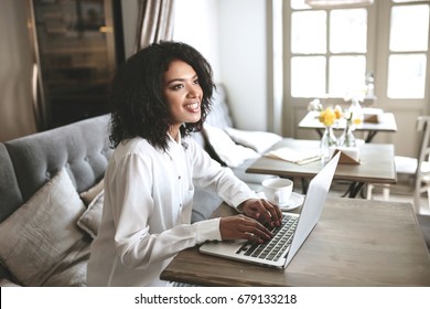 Beautiful Lady Working On Laptop In Restaurant. Pretty African American Girl Typing On Computer In Cafe. Portrait Of Smiling Girl With Dark Curly Hair In White Shirt Sitting In Restaurant