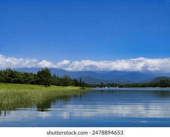 A beautiful lake with clear blue water, surrounded by lush green trees and distant mountains under a vibrant blue sky with scattered clouds, creating a serene natural setting. - Powered by Shutterstock