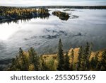 A beautiful lake with trees in the background. The water is calm and the sky is clear. The trees are in various stages of autumn, with some still green and others with leaves falling