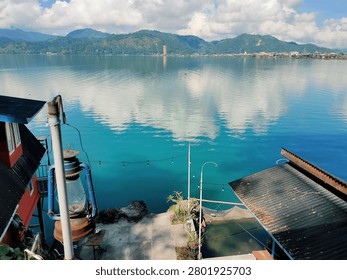 Beautiful lakeside view captured from an elevated waterfront location overlooking crystal-clear blue water and distant green mountains. The lake reflects towering white clouds and the surrounding landscape, creating a calm and inviting atmosphere. In the foreground, a rustic lantern, waterside buildings, string lights, and a small dock area add local character and charm. The combination of natural scenery and human elements makes this image ideal for travel, tourism, hospitality, waterfront living, and lifestyle-related projects.