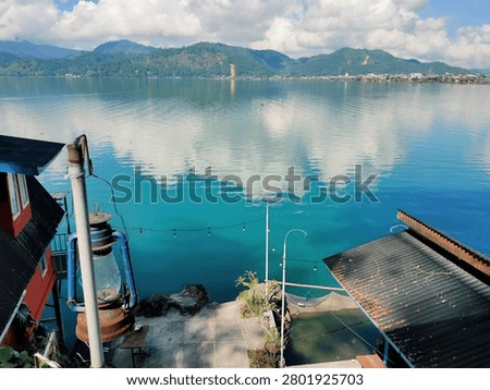 Beautiful lakeside view captured from an elevated waterfront location overlooking crystal-clear blue water and distant green mountains. The lake reflects towering white clouds and the surrounding landscape, creating a calm and inviting atmosphere. In the foreground, a rustic lantern, waterside buildings, string lights, and a small dock area add local character and charm. The combination of natural scenery and human elements makes this image ideal for travel, tourism, hospitality, waterfront living, and lifestyle-related projects.