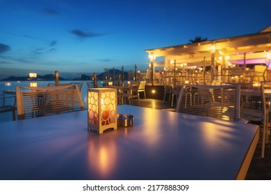 Beautiful lamp on the table in cafe on sea coast at night in summer. Landscape with candle and blurred background with chairs and tables, lights. Empty cozy cafe outdoor with illumination. Travel - Powered by Shutterstock