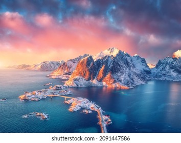 Schöne Landschaft mit blauem Meer, schneebedeckten Bergen, Felsen und Inseln, Dorf, Ror, Straße, Brücke und rosa Himmel bei Sonnenaufgang. Luftbild. Hamnoy in Schnee im Winter auf den Lofoten Inseln, Norwegen. Draufsicht