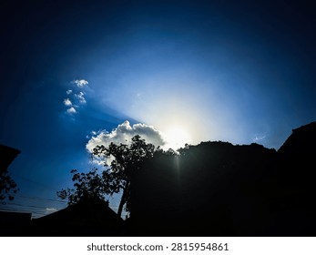 Beautiful landscape featuring dramatic sun rays shining through clouds above silhouetted trees and rooftops under a deep blue sky. Perfect for nature, weather, travel, outdoor, and inspirational themes.
