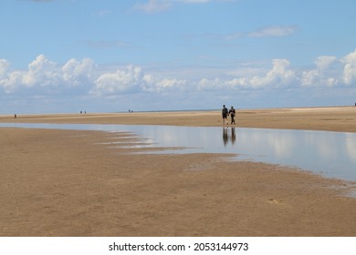 Beautiful Landscape Of Holkham Norfolk Uk Sandy Beach With Couple On Holiday Walk By Ocean Shore With White Puffy Clouds Reflected In Sea Water Lakes Formed On The Sand In Low Tide Summer Blue Skies