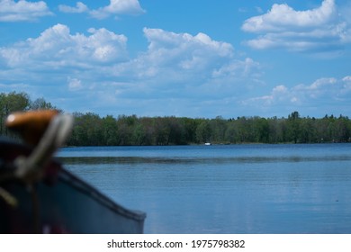 Beautiful Landscape Of Lake.  Green Tree Line Aqua Blue Lake Water And Fluffy Clouds In Blue Sky.  Canoe Out Of Focus In Foreground With Canoe Paddle.  Boat.  Cottage.