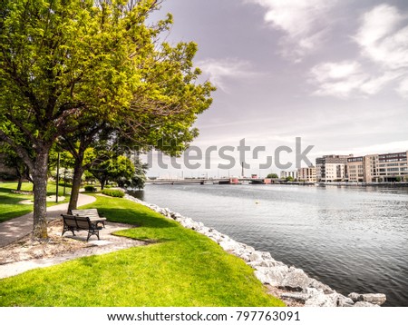 Beautiful landscape scenery shot along the Fox River just after sunrise in Green Bay Wiscosin with riverwalk trail, bench, trees, grass and rocky shoreline framing the shot and blue cloud filled sky.