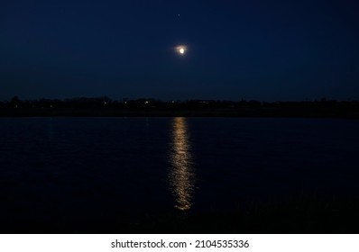 Beautiful Landscape View Of A Glowing Moon Beginning Its Rise Over Lake. A Pale Yellow Aura Surrounds The Moon And A Strip Of Light Reflects On The Water.