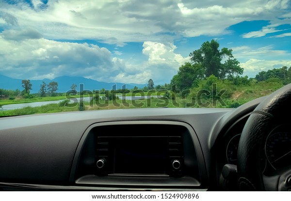Beautiful landscape view from inside car. Steering\
wheel and dashboard of car interior. Road trip travel with scenic\
view of mountain, lake, and forest. Blue sky and white fluffy\
clouds. Vacation time