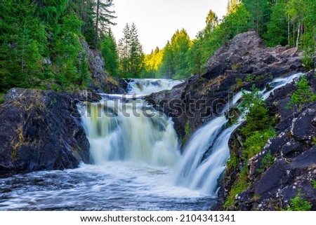 Beautiful landscape with waterfall in northern forest on summer evening. Powerful stream of water among stone rocks and green foliage. Kivach waterfall at Suna river in Karelia, Russia.