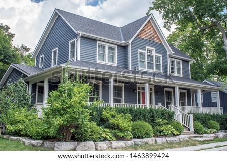 A beautiful large modern custom executive residential house with grey siding and gray shingles and white trim, large over sized windows, lush gardens and green lawn.