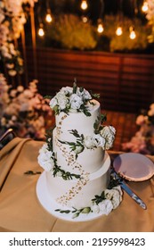 A Beautiful Large Three-tier White, Decorated With Rose Flowers, Wedding Creamy Sweet Cake Stands On The Table In The Evening. Food Photography.