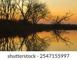 Beautiful late-Autumn landscape captured at Horicon Marsh in Dodge County, Wisconsin, as the sun sets on a peaceful river reflecting silhouetted trees beneath an orange sky.