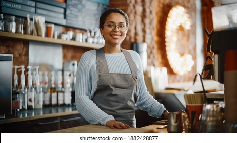 Beautiful Latin American Female Barista With Short Hair And Glasses Is Projecting A Happy Smile In Coffee Shop Bar. Portrait Of Happy Employee Behind Cozy Loft-Style Cafe Counter In Restaurant.