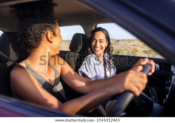 Beautiful\
latin hispanic woman with friend enjoying and laughing in car while\
going on a road trip. Cheerful girl friends enjoying trip and drive\
in car at sunset. Black woman driving a\
car.