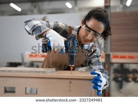 Beautiful Latin woman carpenter using power tools working her wood job in carpenter's shop. Young hispanic female in protective goggles busy in furniture woodworking. Feminism in carpentry industry.