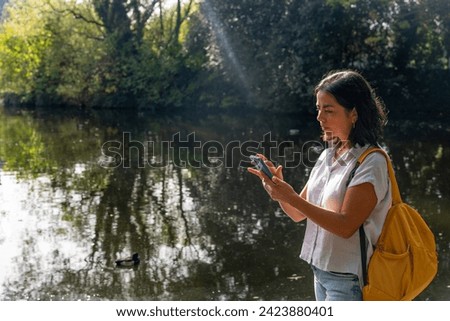 Beautiful latin woman checking her cell phone next to a pond in a Dublin park with a ray of sunlight illuminating her