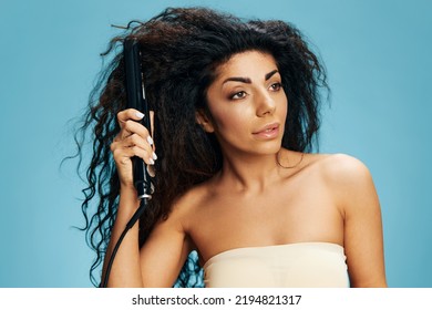 Beautiful Latin Woman With Curly Afro Using Straightener, Looking Aside, Posing Isolated On Blue Wall Background. Dry Damaged Hair Routine Concept, Haircare, Ironing, Hairdressing