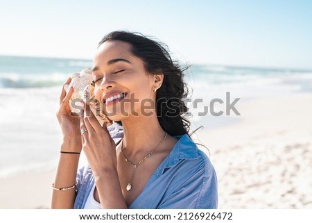 Beautiful latin woman hearing the sound of the sea with a big seashell at beach. Hispanic girl on seacoast with a cockleshell in hands with copy space. Woman on a white sand beach holding a sea shell.