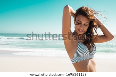 Beautiful latin woman in striped bikini on tropical beach with copy space. Stylish and fashion young woman smiling and looking down at sea. Tanned girl in swimwear enjoying and walking on beach.