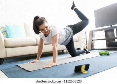 Beautiful Latin Young Woman Repeating Exercises While Watching Online Exercise Session On Her Smart Phone At Home