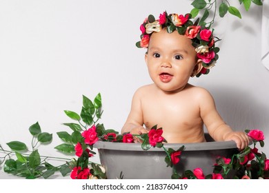 Beautiful Latina Baby Girl With Brown Skin, Very Smiling And Happy With A Crown Of Flowers In A Bucket Surrounded By Flowers. Seated Girl. White Background With Space To Copy Text
